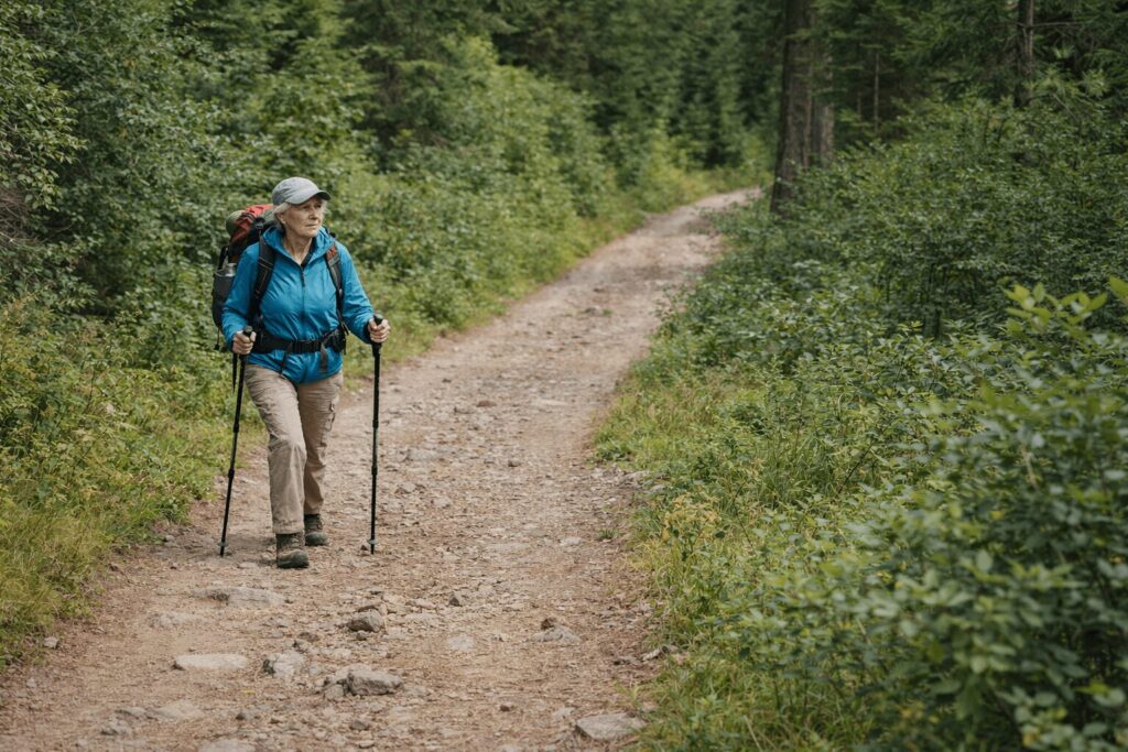Older woman hiking alone on a forest trail with trekking poles and a backpack, illustrating deliberate solo travel and personal responsibility.