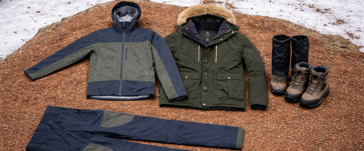 Outdoor shell layer clothing system laid out on a forest floor, showing waterproof jackets, horizontal shell pants, hiking boots, and boot gaiters arranged clearly for weather protection.