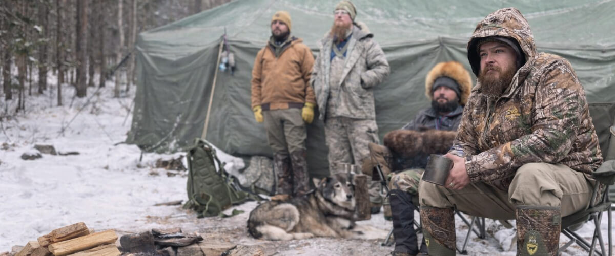 Men wearing layered cold weather clothing sitting around a winter campsite fire with a canvas tent and snow-covered forest in the background