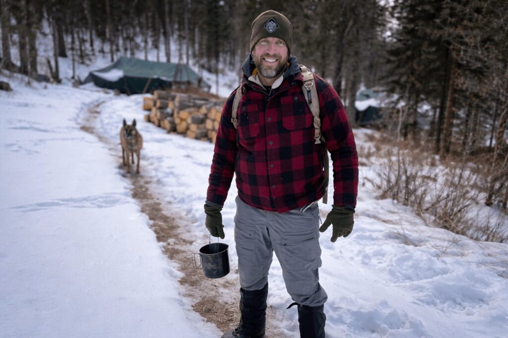 Man dressed in winter layers standing on a snowy forest trail with backpack, gloves, insulated pants, winter boots, and a dog following behind in a cold weather camp setting.