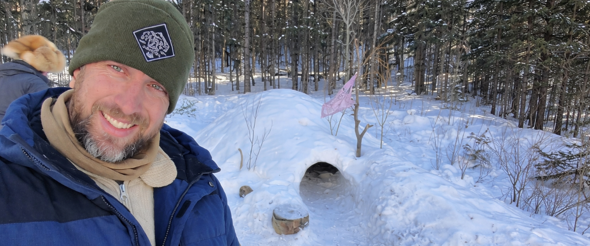 Man wearing winter clothing layers standing beside a snow shelter in a forest, demonstrating practical cold weather layering in real outdoor conditions.
