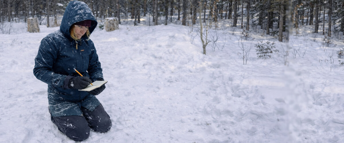 Woman kneeling in a snowy forest adjusting winter clothing layers during cold weather outdoor training.