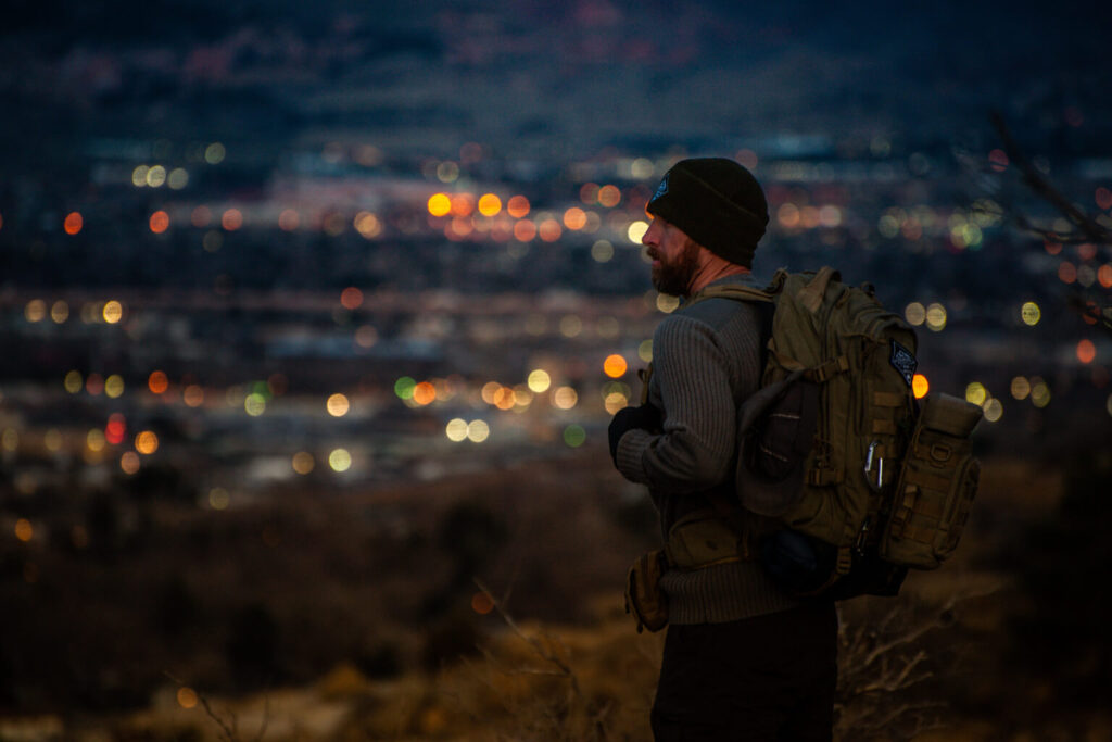 A person wearing a backpack stands on a hillside at night overlooking a city filled with glowing lights, observing the urban landscape from above in quiet reflection