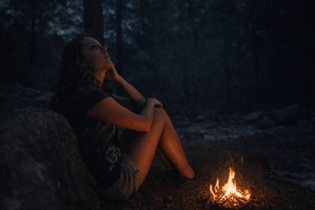 A woman sits alone in a dark forest at night beside a small ground fire, illuminated by its glow as rain falls and conditions shift around her