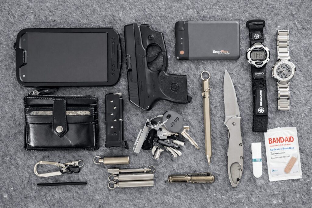 Everyday carry EDC flat lay showing concealed carry pistol, spare magazine, folding knife, flashlight, wallet, keys, power bank, watches, multitool, and basic first aid items arranged on gray surface.
