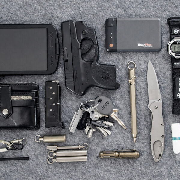 Everyday carry EDC gear layout including concealed carry pistol, spare magazine, folding knife, flashlight, wallet, keys, power bank, watch, and multitool arranged on gray fabric surface.