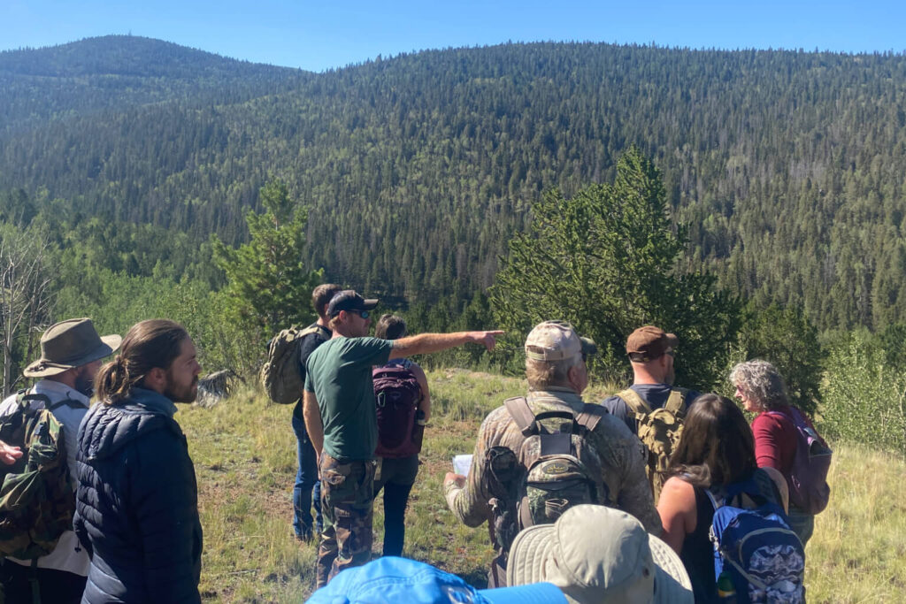 Group of backpackers paused on an open hillside while one person points toward distant terrain, with the rest of the group turned toward him and listening during a navigation discussion.