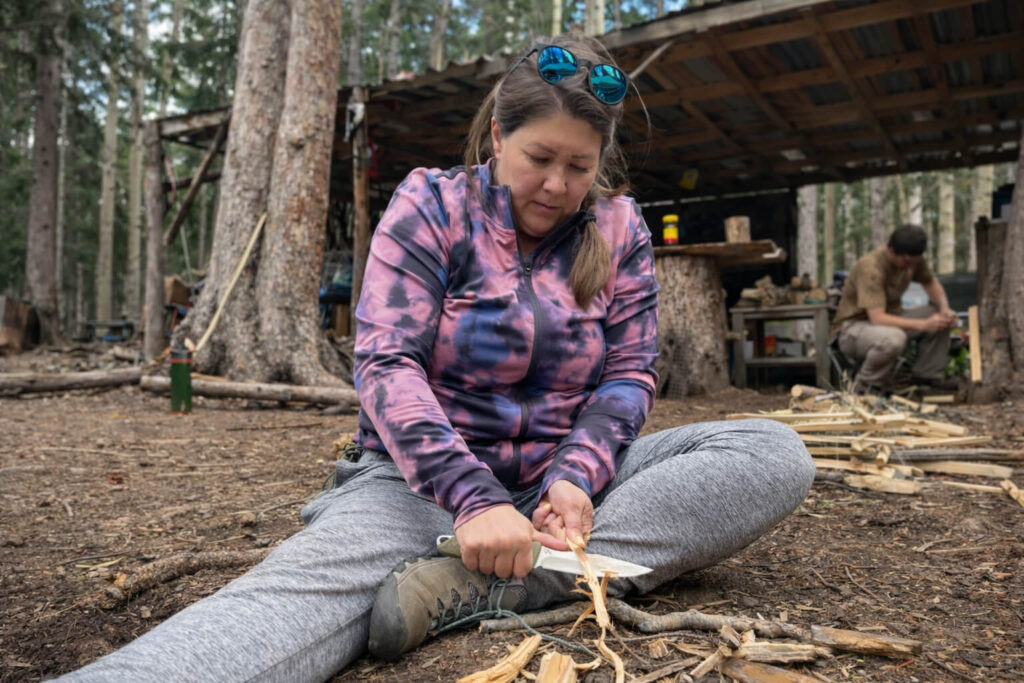 A woman sits on the forest floor carving wood with a knife while practicing a skill in an outdoor learning environment with a simple structure behind her