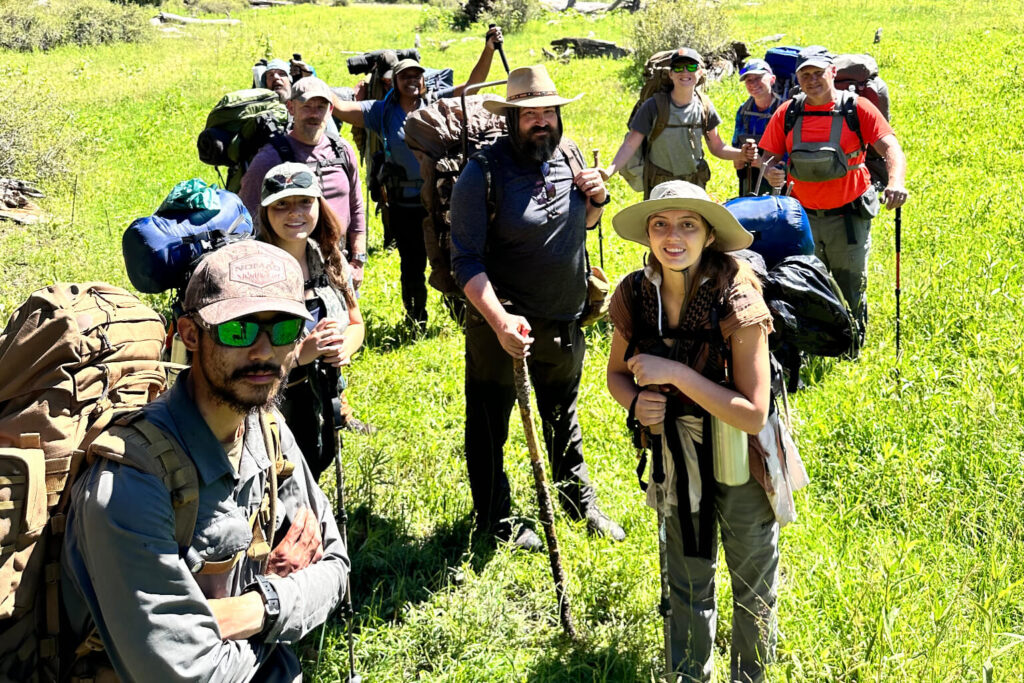 Group of backpackers standing close together in a grassy mountain meadow, wearing packs and holding trekking poles, with forested hills and clear blue sky in the background, showing a cohesive group traveling together.