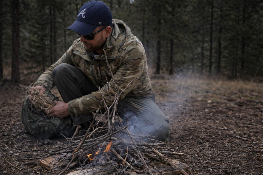 Person tending a weak, smoky fire under marginal conditions during outdoor skills training