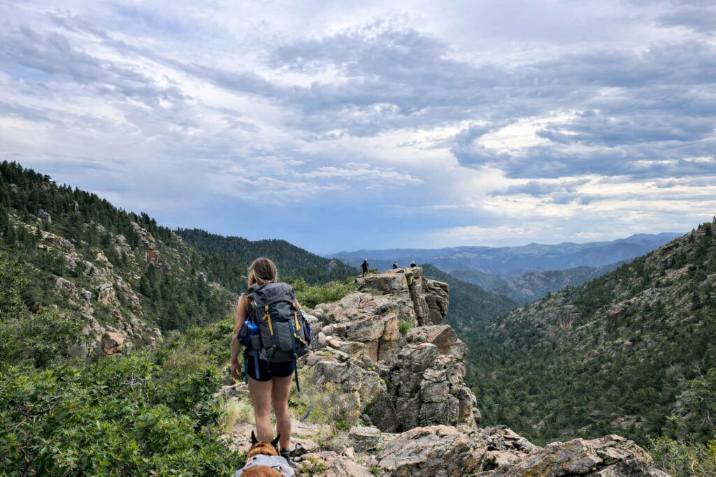 A hiker with a smaller backpack walks along a rocky mountain ridge with her dog while distant hikers stand on an outcrop overlooking a forested valley and layered mountain ranges under a cloudy sky