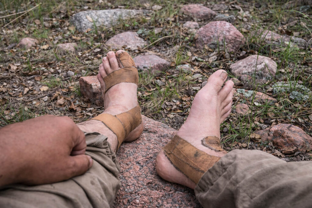 Taped bare feet resting on pink granite rocks during a backcountry hike, showing worn athletic tape, dirt, and early foot damage with grass, leaves, and scattered stones on the forest floor.