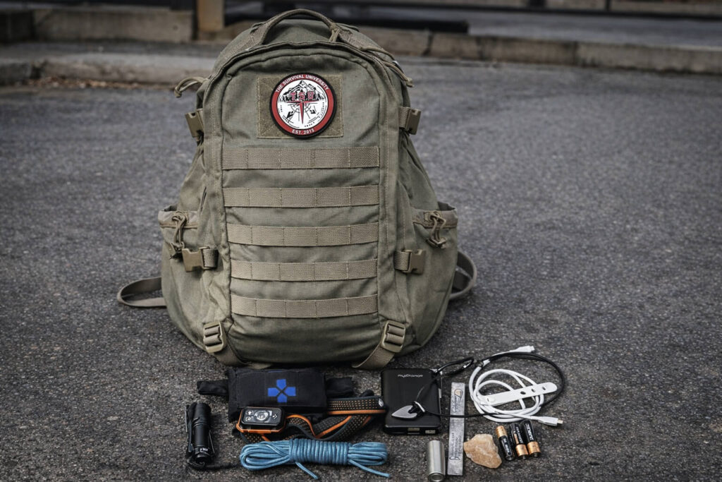 Everyday carry backpack placed on city pavement with organized EDC gear including flashlight, headlamp, paracord, power bank, charging cables, batteries, fire starter, and compact essentials arranged in front.
