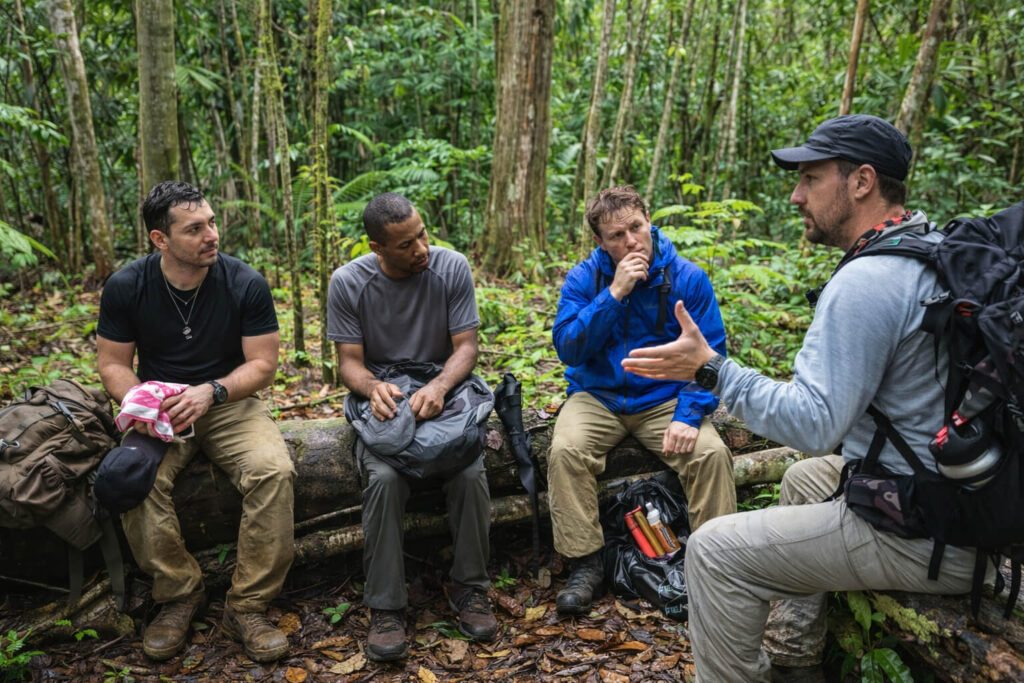Group of backpackers paused in a Costa Rica jungle, sitting on a fallen log while one person on the right speaks and gestures, with the others turned toward him and listening during a regroup in dense tropical forest.