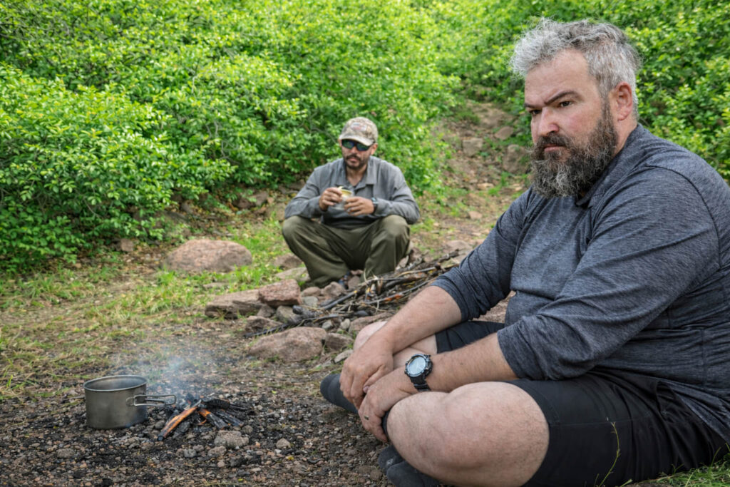 Two men resting quietly at a forest campsite during daylight, showing the slow and unglamorous reality of outdoor survival