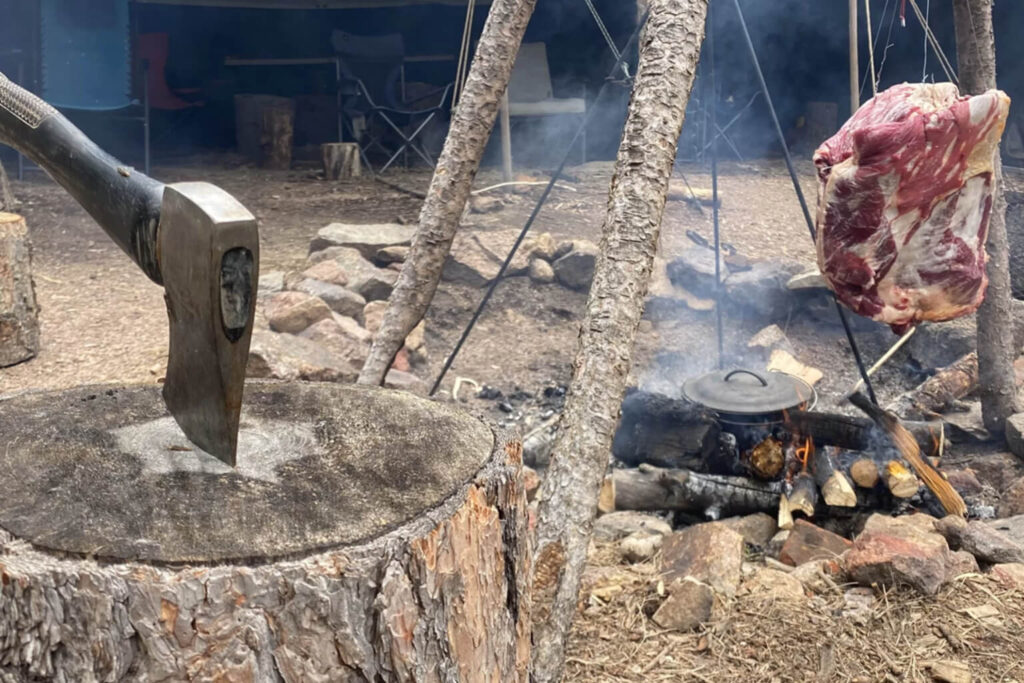 Large cut of meat hanging beside a campfire tripod with a cooking pot and an axe embedded in a chopping block at a wilderness camp