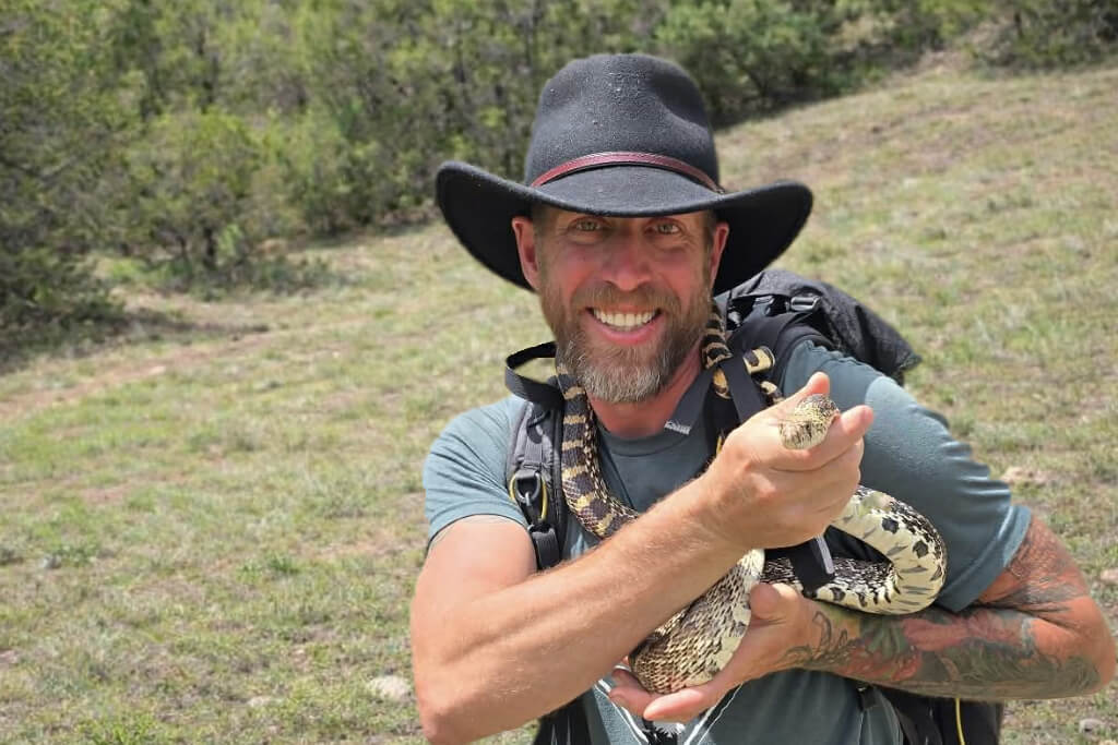 Jason Marsteiner holding a Bullsnake in a grassy Colorado hillside environment, demonstrating snake handling and identification in the field