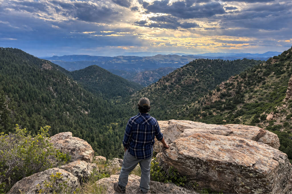 Hiker standing on a rocky overlook studying the surrounding mountain terrain and valleys to maintain awareness and stay oriented while hiking