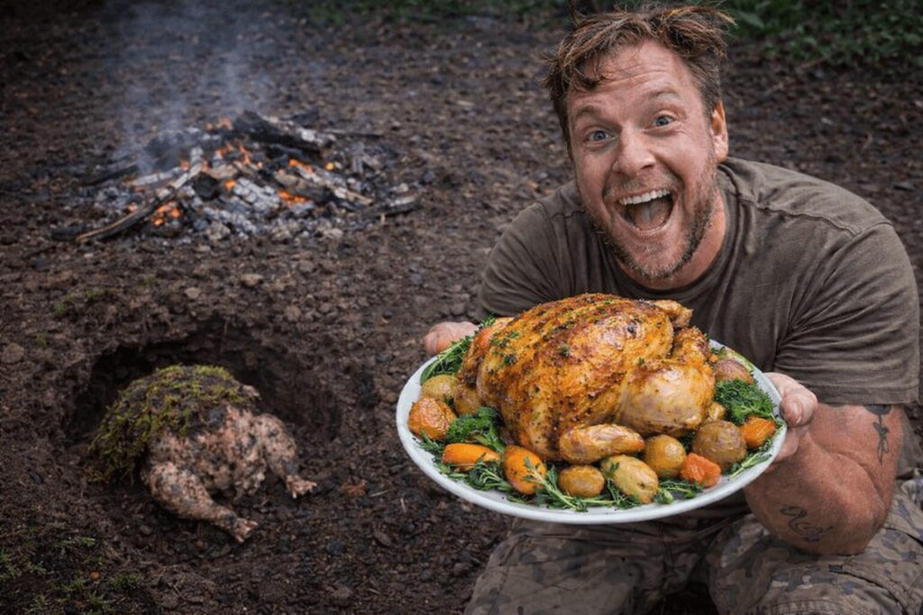 Man holding a perfectly roasted chicken on a plate while a raw dirty chicken covered in moss sits in a hole in the ground behind him next to a smoldering fire
