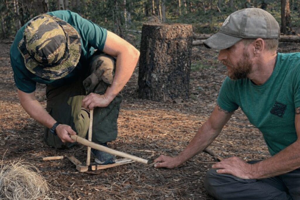 Instructor teaching a student how to start a fire using a traditional bow drill in the forest