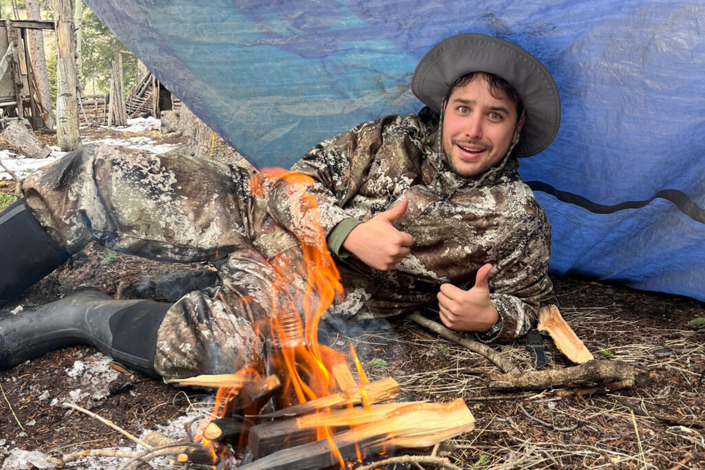 Person staying warm beside a small fire under a tarp shelter in cold wilderness conditions