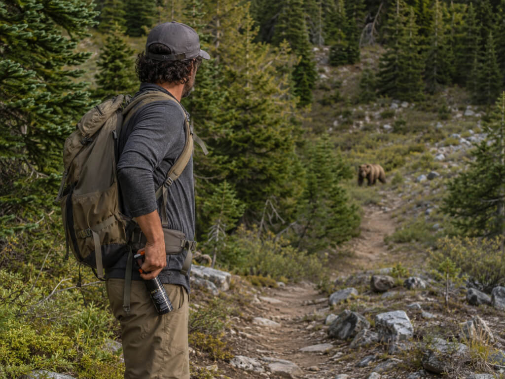 Hiker holding bear spray on a mountain trail while a brown bear stands at a safe distance in the background.
