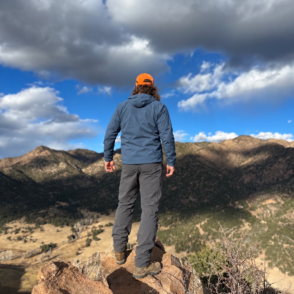 Hiker in a blue jacket and orange cap standing on a rocky overlook above a mountain valley beneath dark clouds.