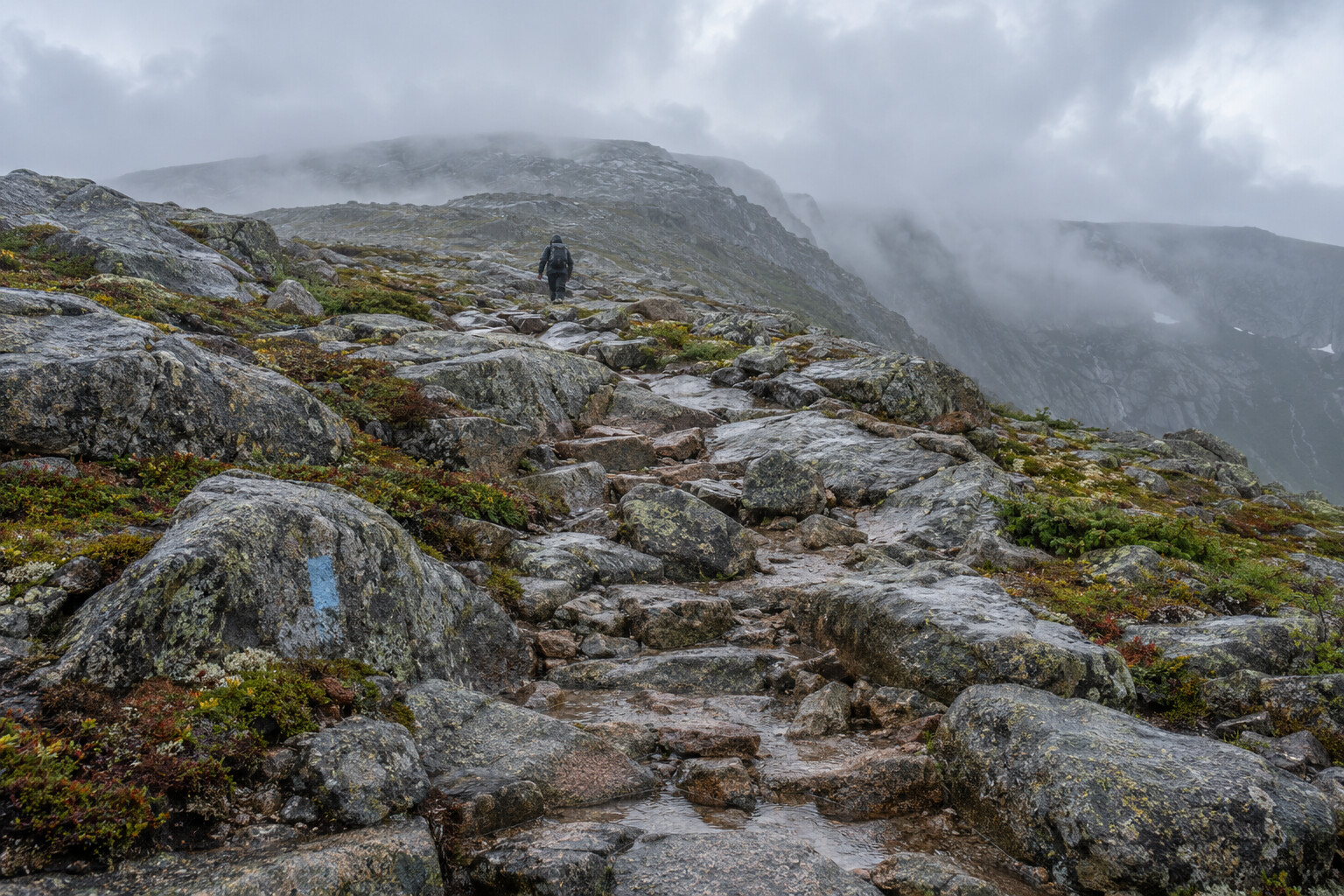Wet granite boulders and a distant hiker on an exposed alpine route in low cloud.
