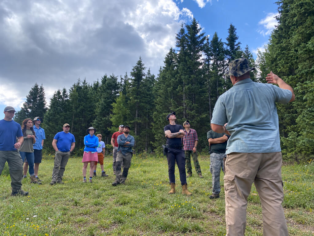 Survival instructor addressing a group of students in a grassy clearing surrounded by evergreen trees.