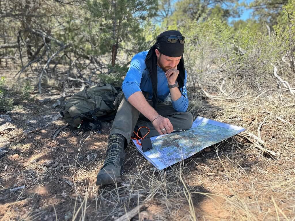 Hiker seated in a pine forest studying a topographic map spread on the ground beside his backpack.