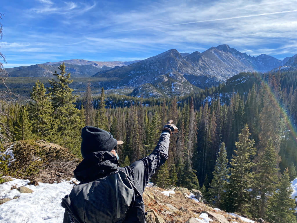 Hiker with a backpack pointing toward rugged mountain peaks from a snowy overlook above an evergreen forest.