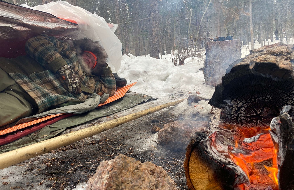 Person resting on an insulating sleeping pad beneath a shelter beside a campfire in a snowy forest.