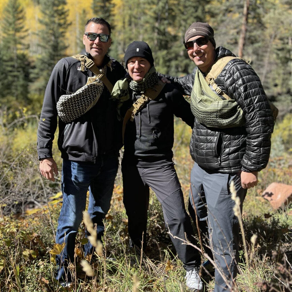 Three men in outdoor gear pose together in a forest clearing surrounded by tall grass and colorful autumn trees.